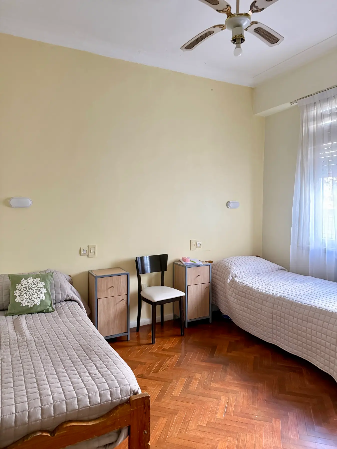 Habitación compartida con dos camas, mesas de luz y piso de parquet, iluminada por luz natural