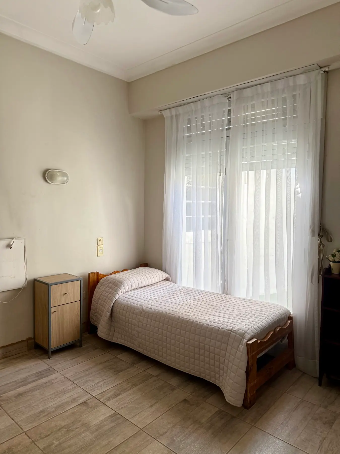 Habitación individual con cama, mesa de luz y ventanal con cortinas al balcón
