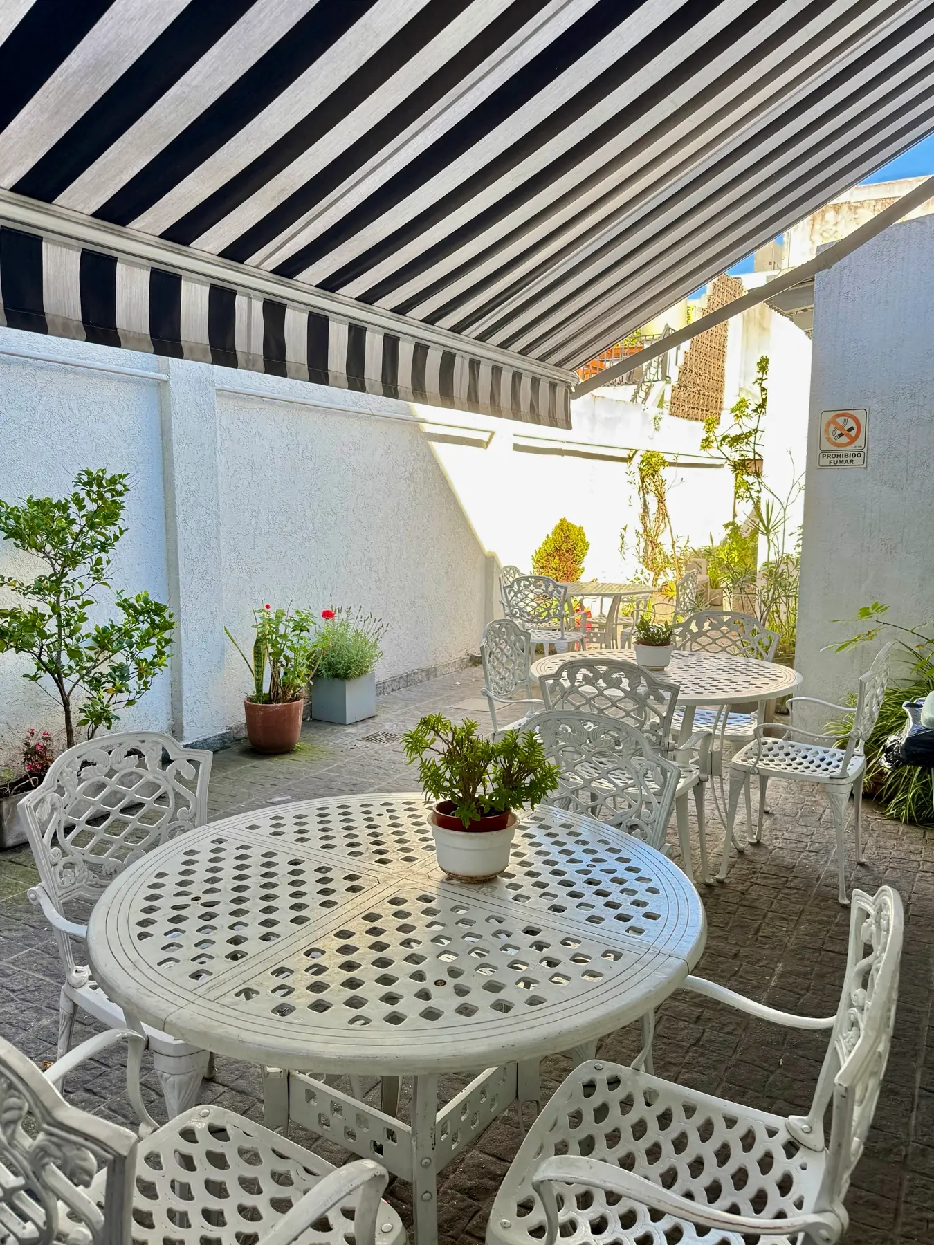 Galería exterior con toldo rayado, mesas de hierro blanco y plantas en macetas