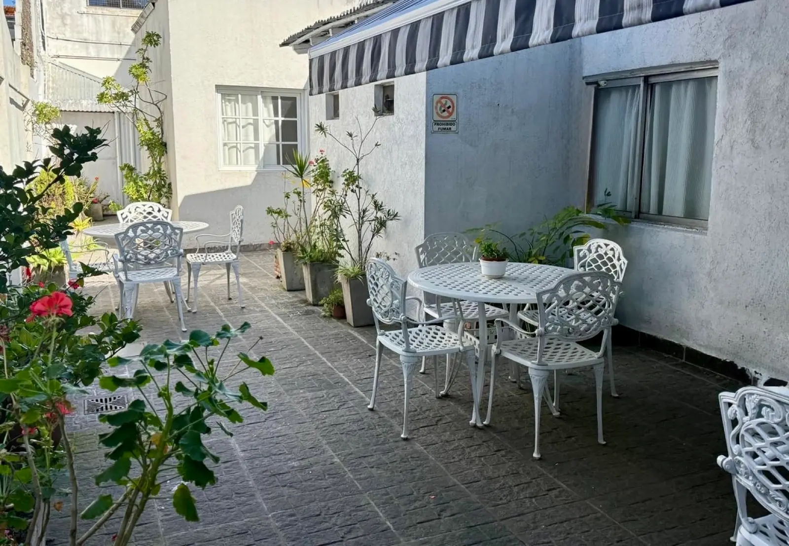 Patio exterior con varias mesas y sillas de hierro blanco entre canteros con flores