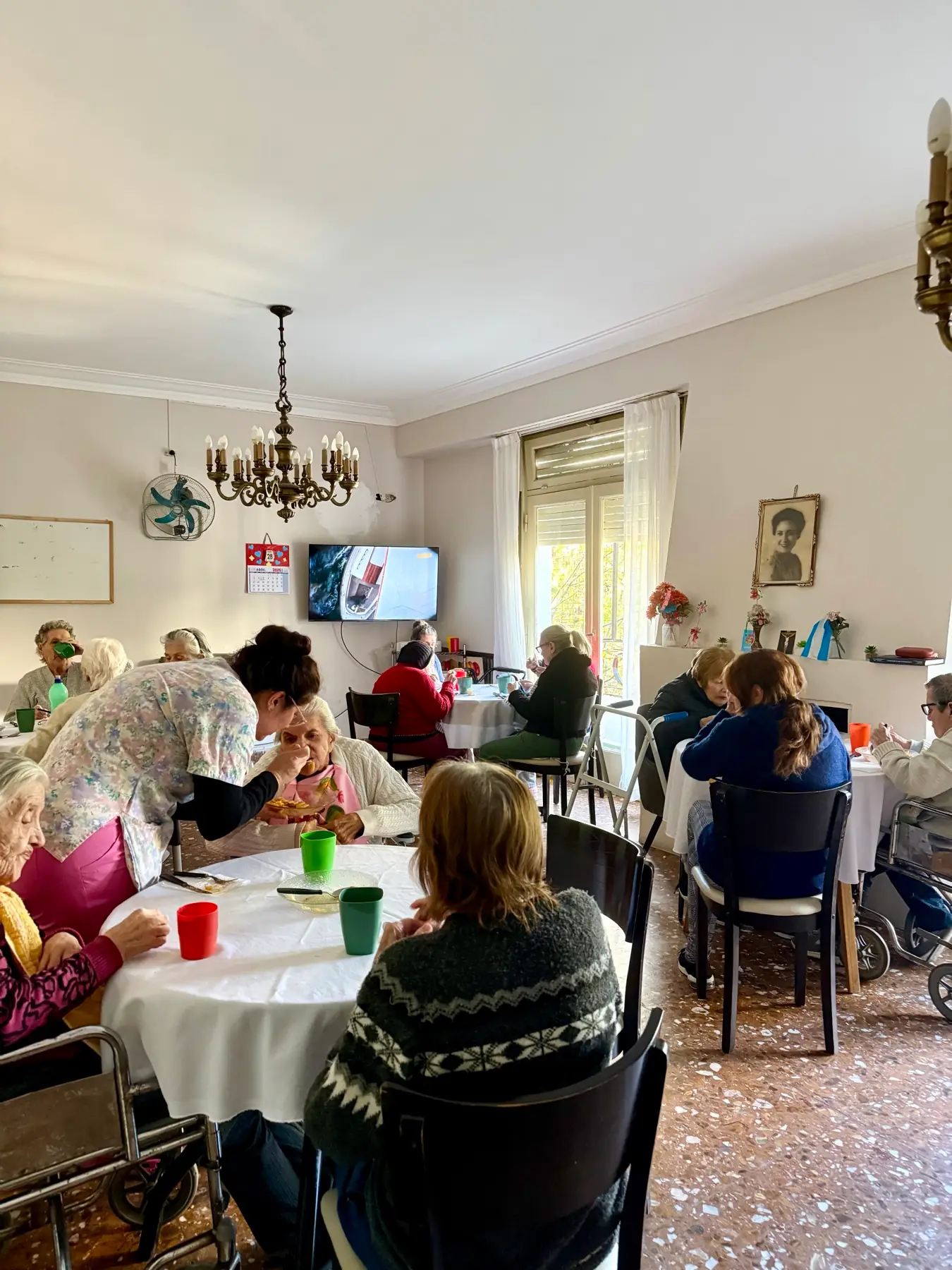 Vista del salón comedor con residentes en las mesas y el televisor encendido